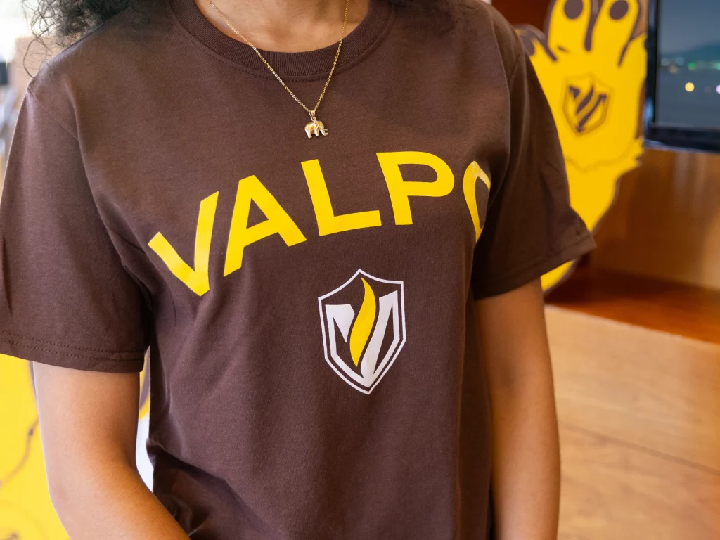 Person wearing a brown "VALPO" t-shirt with a yellow and white shield logo from Our Logos, paired with an elephant necklace.