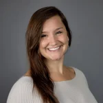 Katie Zromkoski, OTD, a woman with long brown hair smiling and wearing a light gray top, stands against a plain gray background.