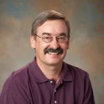 Man with glasses and a mustache smiling, wearing a purple polo shirt, in front of a neutral background.