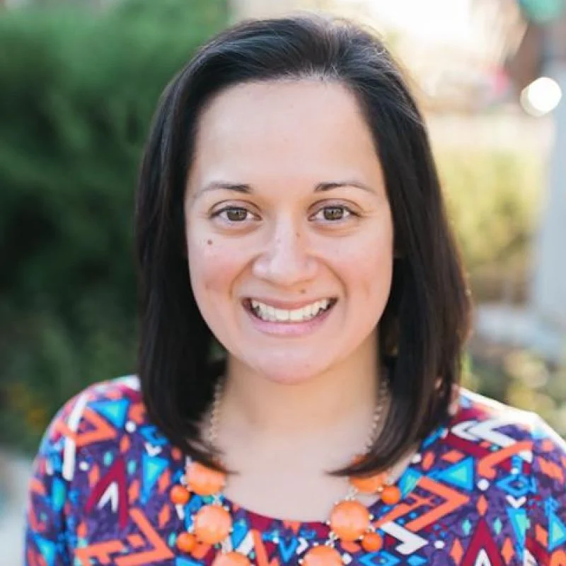 Woman with shoulder-length dark hair, smiling, wearing a colorful patterned top and orange necklace outdoors.