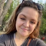Zoe Vander Vinne, with wavy hair and sunglasses on her head, smiles outdoors with trees in the background.