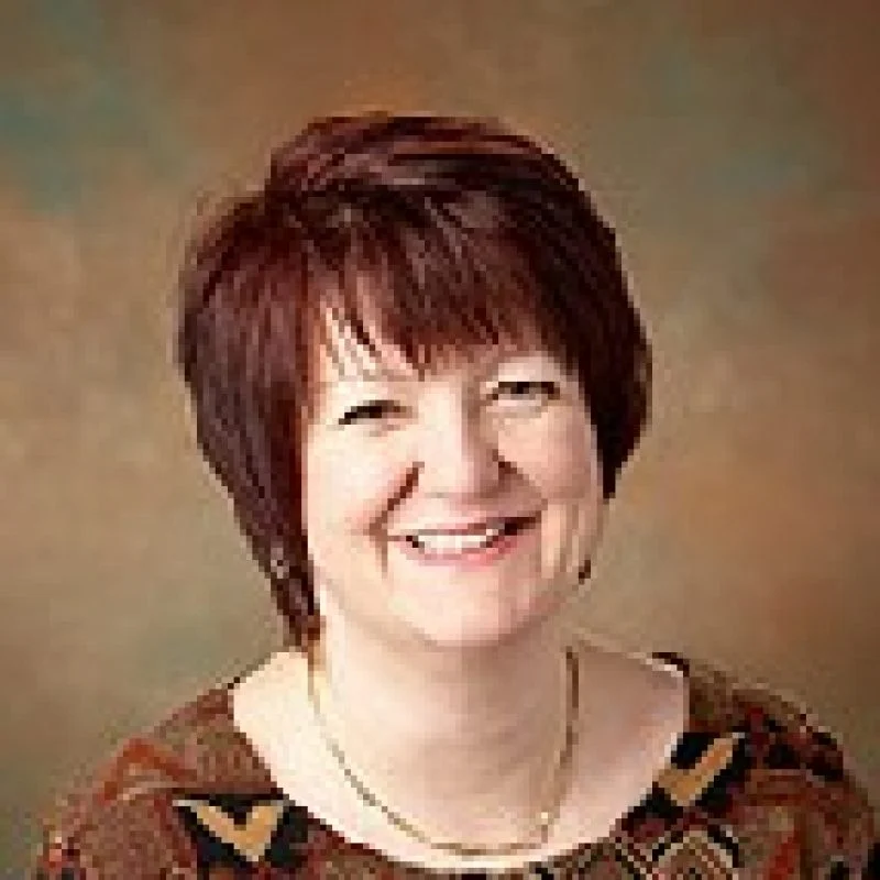 Smiling woman with short brown hair wearing a patterned top, in front of a brown background. Zsuzsanna Szaniszlo.